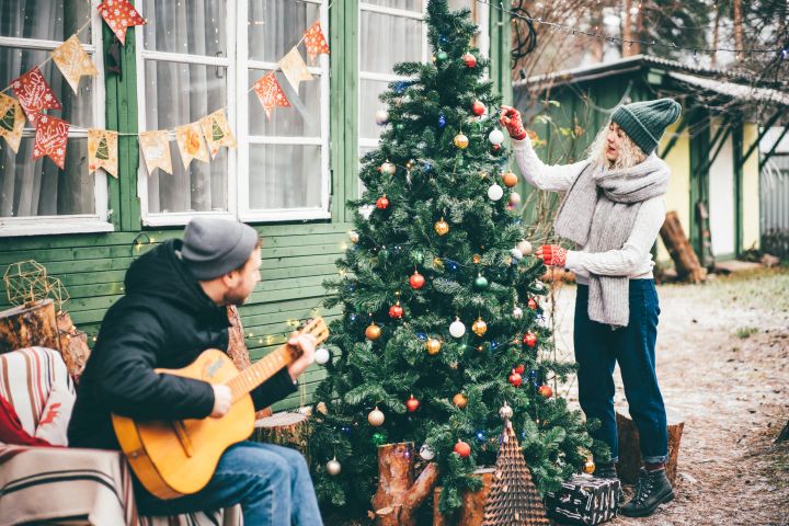Frau schmückt Weihnachtsbaum vor Haus, Mann sitzt daneben und spielt Gitarre