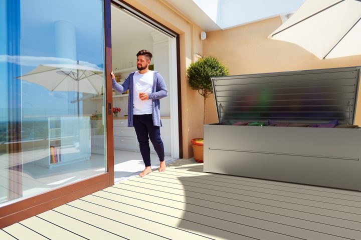 Ilesto Auflagenbox aus Stahl in silber steht auf einem Balkon, daneben steht ein Mann in der Türe und genießt die Sonne