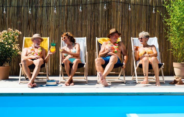 Poolparty: vier ältere Menschen sitzen mit Cocktails in der Hand in Liegestühlen am Pool