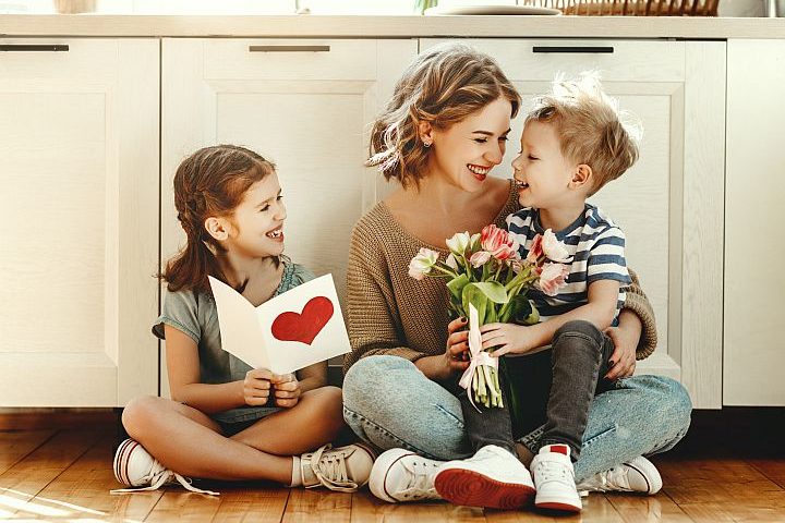 Mutter sitzt mit ihren zwei Kindern in der Küche am Boden, Sohn sitzt auf ihrem Schoß und überreicht ihr einen Blumenstrauß zum Muttertag, Tochter sitzt daneben und überreicht ihr eine Muttertagskarte