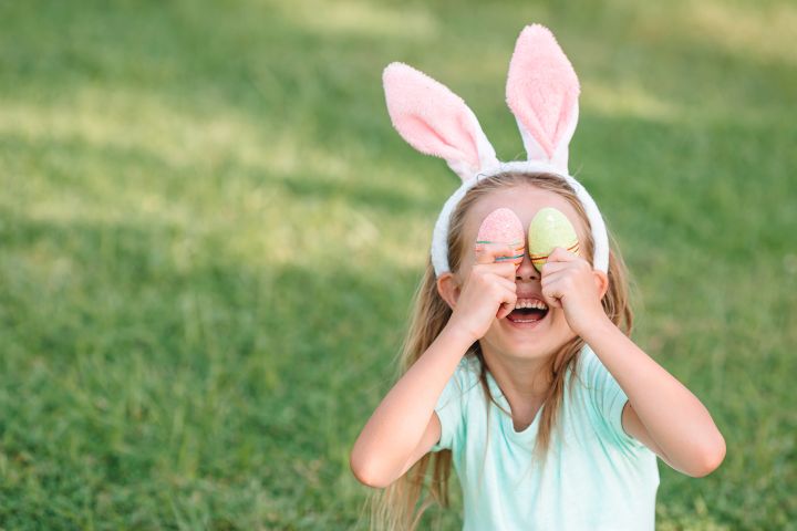 Mädchen mit Hasenohren hält zwei Ostereier vor ihre Augen und lacht in die Kamera, im Hintergrund grüne Wiese