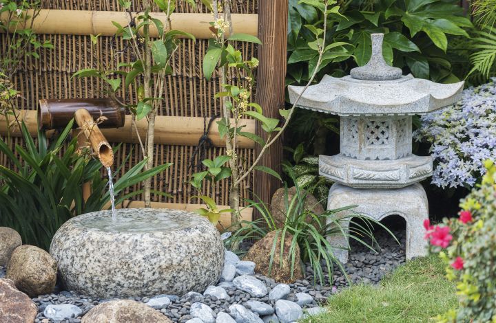 Man sieht eine Steinlaterne neben einem kleinen Brunnen, gebaut aus Bambusstielen, darunter ein großer Stein auf den das Wasser rinnt