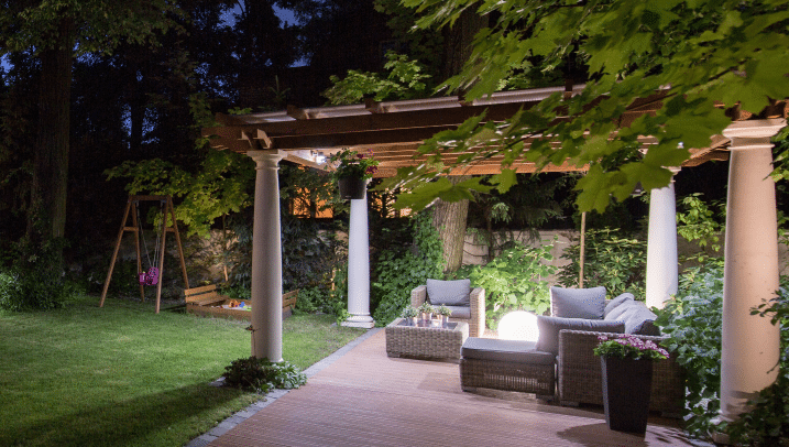 Gartenpavillon in der Nacht mit weißen Säulen und Holzdach, darunter Sofas und ein Tisch, rechts eine Schaukel und ein Sandkasten