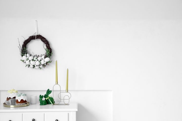 Tisch mit Osterkuchen, Osterhasen, Kerzen nah einer weißen Wand, auf der ein Osterzweig mit weißen Blumen hängt