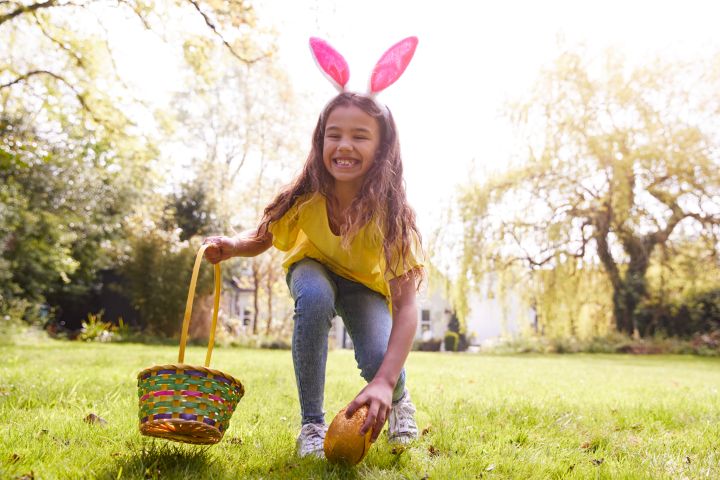Ein Mädchen sucht nach Osternestern und greift gerade lachend zu einer Osterpinze in der Wiese
