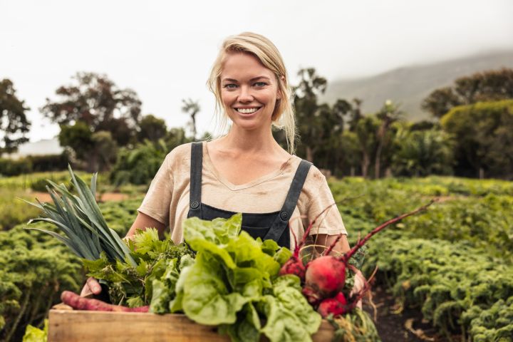 Bäuerin lächelt in die Kamera, in den Armen hält sie eine Kiste voller frisch geerntetem Gemüse