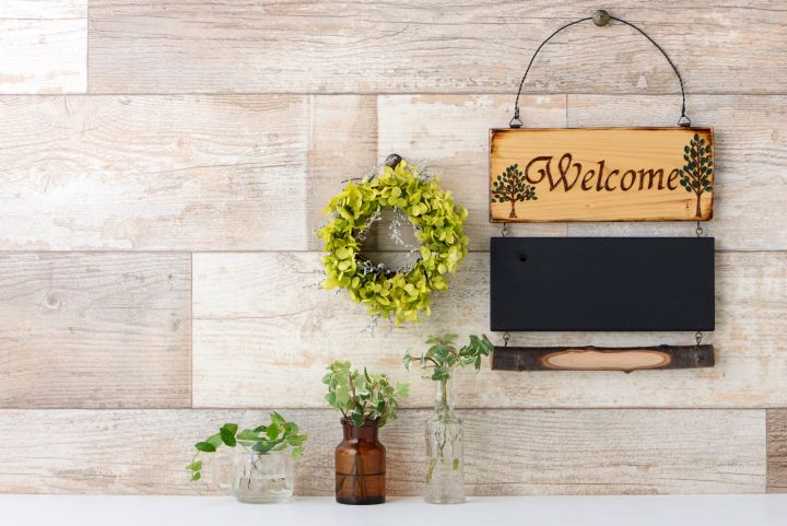 Vintage-Schild mit der Aufschrift Welcome aus Holz hängt an einer Holzwand. Daneben ein Kranz und darunter Blumenvasen auf einem Tisch.